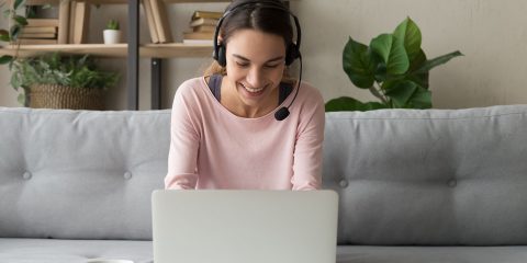 Smiling young woman student wear wireless headset using laptop for e learning course online teaching in internet, happy female teacher tutor communicating by video conference call sit on sofa at home