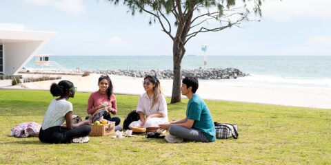 Four students sitting on grass with a picnic. Background shows the ocean and sunny sky
